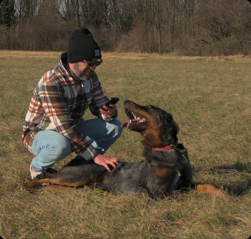 Uomo moro accovacciato consulta smartphone in compagnia di beauceron arlecchino che lo guarda