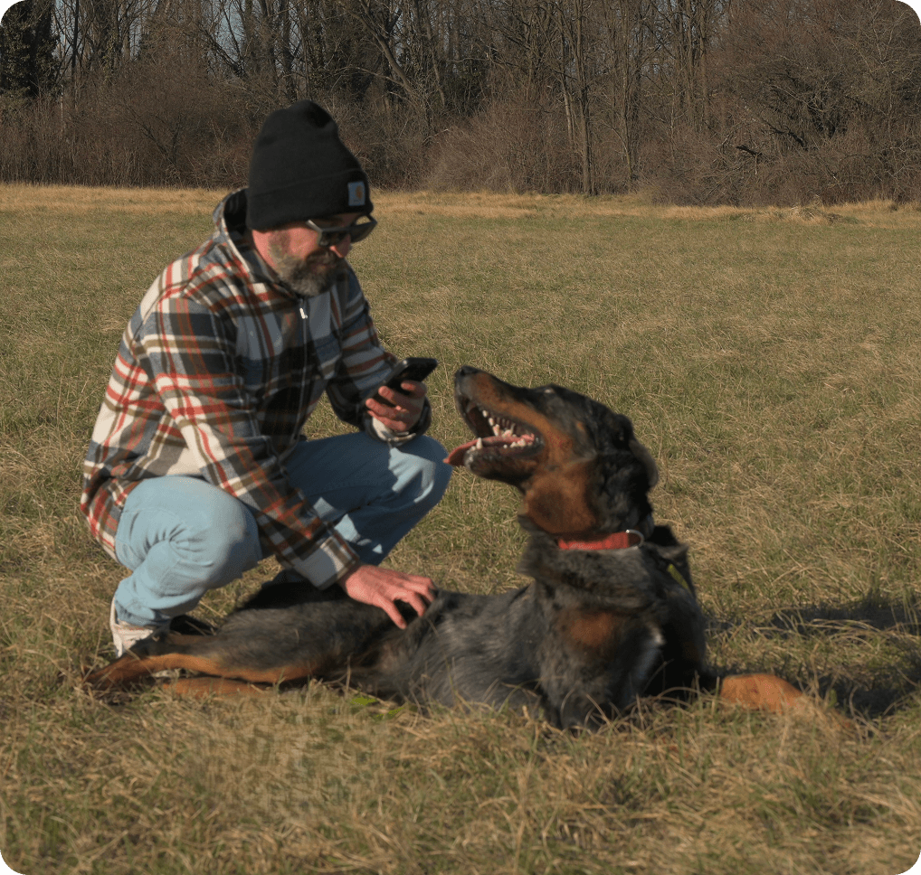 Uomo moro accovacciato consulta smartphone in compagnia di beauceron arlecchino che lo guarda